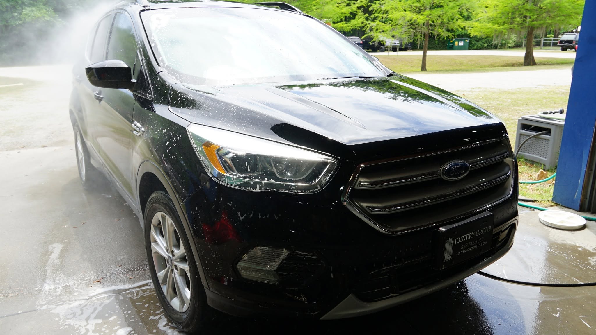 Black Ford SUV during an exterior wash, with soap on the windshield and water spraying beside it