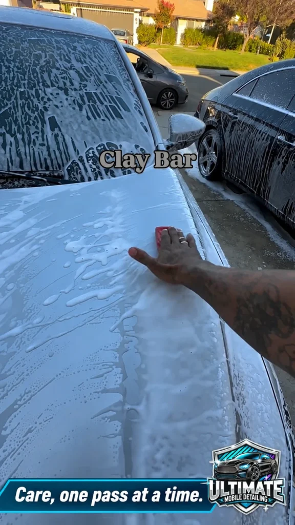 Hand working with a red claying tool near the outer edge of a wet vehicle hood